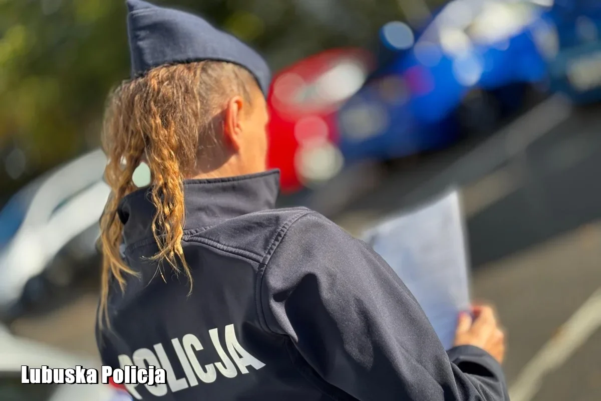 Policjantka w mundurze z napisem „Policja” stoi tyłem na drodze, trzymając dokument, w tle widać policyjny radiowóz.
