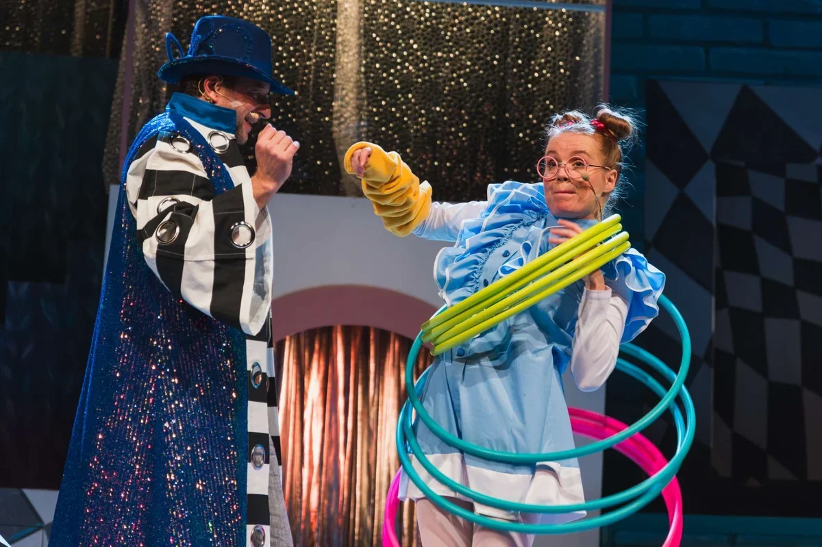 Dwie osoby w kolorowych kostiumach scenicznych, jedna z nich trzyma kilka hula-hopów, podczas przedstawienia teatralnego.
