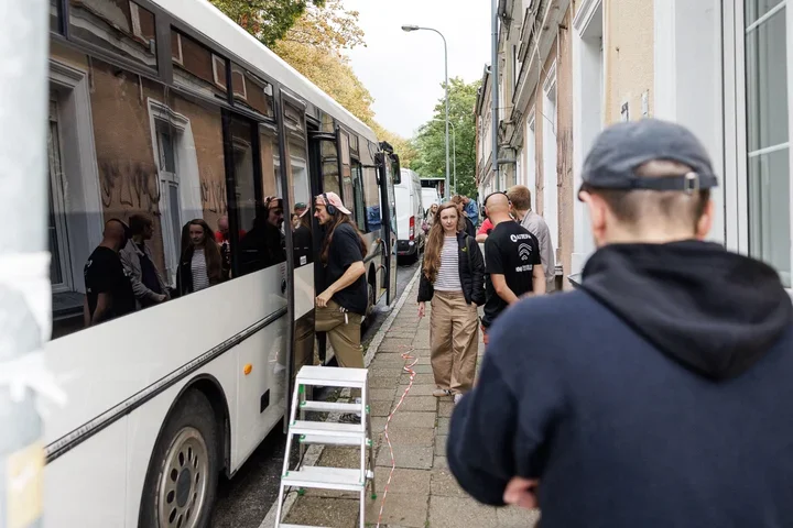 Grupa osób wchodzi i stoi przy białym autokarze zaparkowanym wąskiej ulicy między kamienicami.