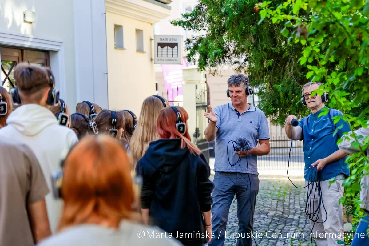 Grupa osób w słuchawkach uczestniczy w plenerowej prezentacji prowadzonej przez dwóch mężczyzn z mikrofonem na brukowanej uliczce przed budynkiem teatru.