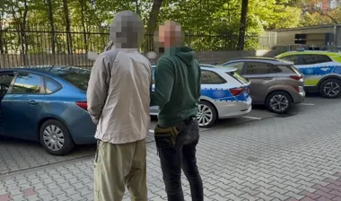 Dwie osoby z zamazanymi twarzami stoją tyłem na parkingu obok policyjnych radiowozów i innych samochodów.