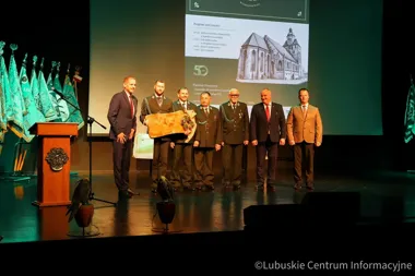 Grupa mężczyzn w garniturach i mundurach stoi na scenie podczas uroczystości jubileuszowej, jeden z nich trzyma drewnianą pamiątkę; w tle ekran z programem obch