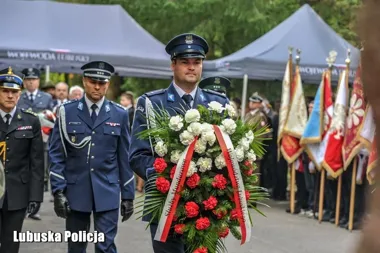 Funkcjonariusze policji w mundurach galowych niosą wieniec z białymi i czerwonymi kwiatami podczas uroczystości z udziałem pocztów sztandarowych.