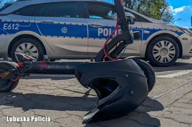 Na brukowanej nawierzchni leży czarny kask i elektryczna hulajnoga, w tle stoi oznakowany radiowóz Lubuskiej Policji.
