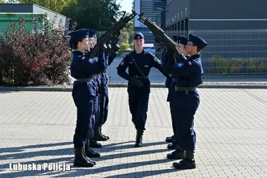 Funkcjonariusze w granatowych mundurach Lubuskiej Policji ćwiczą musztrę z bronią na brukowanym placu przed budynkiem.