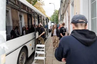 Grupa osób wchodzi i stoi przy białym autokarze zaparkowanym wąskiej ulicy między kamienicami.