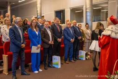 Grupa osób w sali konferencyjnej stoi przed osobą w czerwonym stroju z insygnium, podczas uroczystości z okazji rozpoczęcia roku akademickiego.